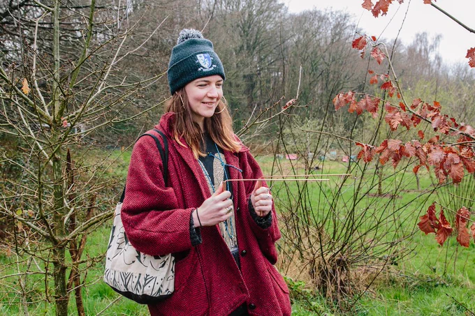 A smiling person wearing a beanie and red coat holding a thin rod, outdoors in nature on a cloudy day.