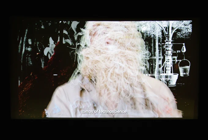 A projection of a person obscured by a mass of string or yarn with a black-and-white backdrop of laboratory equipment, accompanied by the subtitles which read "tombs of technoscience."