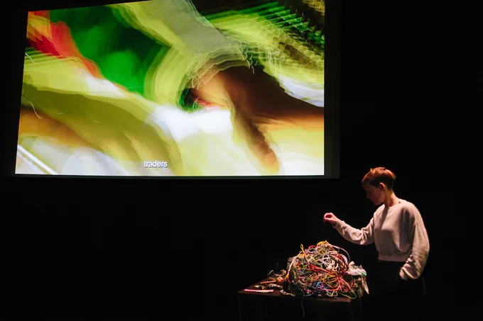 A person interacting with a pile of tangled wires on a table in front of them. Behind them is a colourful, abstract projection.