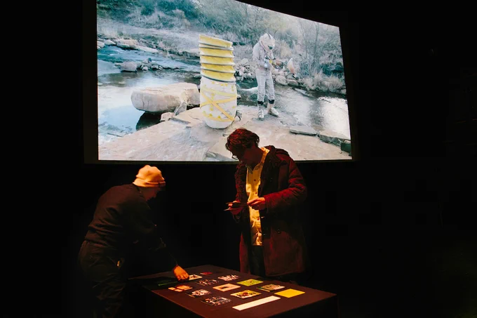 Two people examining postcards on a table with a large projection of an astronaut in a natural landscape in the background.
