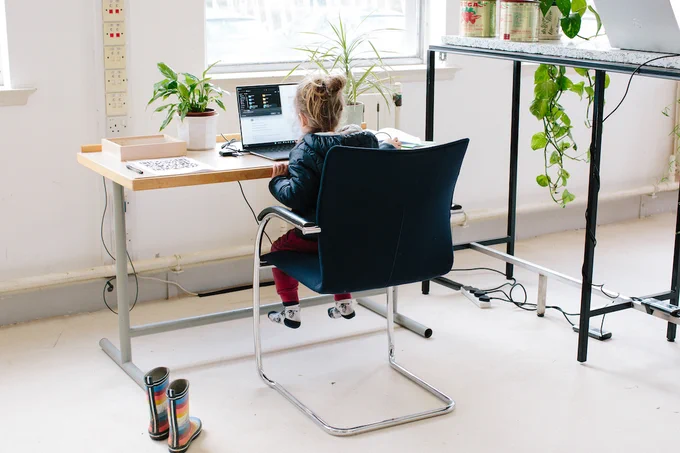 A child sits at a desk using a laptop in a room with large windows and potted plants.