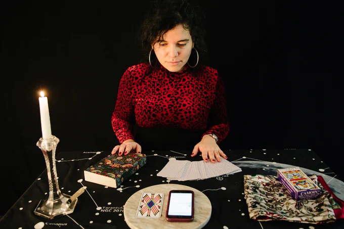 A person sat at a table covered with a candle, book, traditional tarot deck and mobile phone.