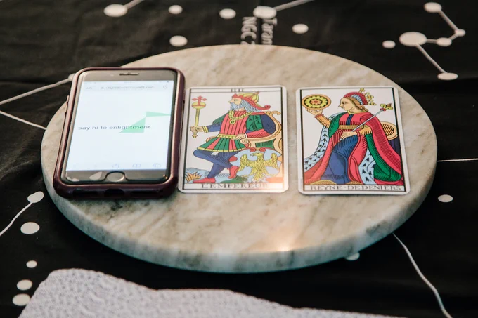 Close-up of a smartphone with the text "say hi to enlightenment" on its screen. Next to it are two tarot cards, 'L'Empereur' and 'Reyne de Deniers'.