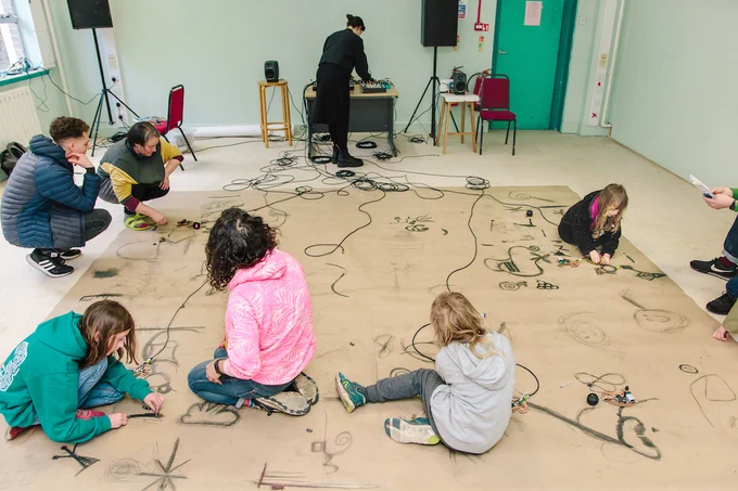 People drawing and wiring electronic circuits on the floor, with audio equipment set up in the background.