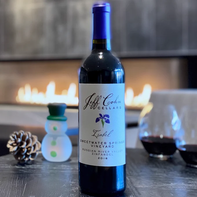 A bottle of 2018 Sweetwater Zindandel Isabel on a table with two glasses of wine on a table with a snowman and fire in the background.