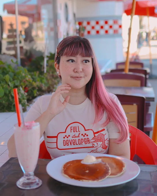 me, a feminine person with pink hair, wearing a shirt that says “full stack developer”, while sitting at an outside table of a diner, eating pancakes and drinking a milkshake