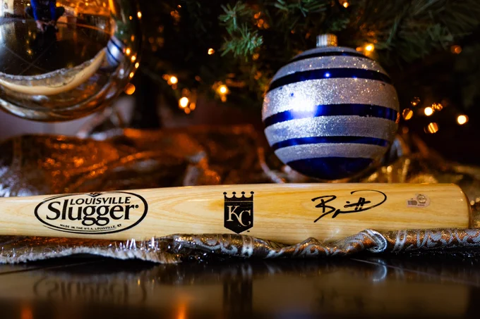 An image of a Louisville Slugger baseball bat signed by Bobby Witt Jr. on the barrel and with a Kansas City Royals logo. The bat is sitting underneath a Christmas tree big ornaments. 