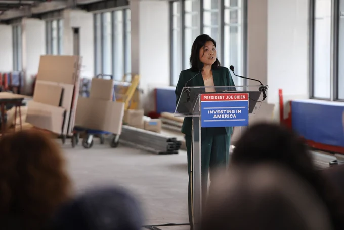 Acting Secretary of Labor Julie Su speaks at a podium at a construction site. The podium has a sign on it that says “President Joe Biden, Investing in America.”