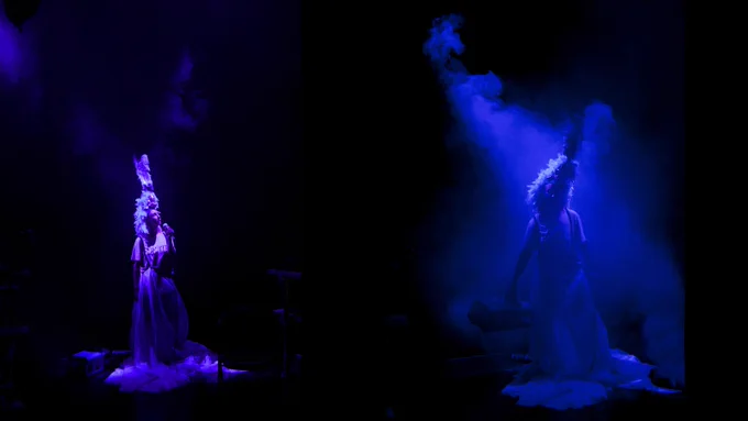 Two photographs of a person standing in the darkness with a purple spotlight shining on them. In one of the photographs, they are surrounded by smoke haze. In both photographs, they are wearing a feathered headdress and a long fabric skirt with a t-shirt which says ‘Death’. Photograph by Holly Revell, design by Eleanor Field. 