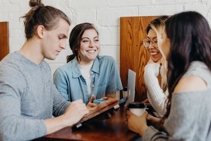 4 students sat around a coffee bar with take out coffee cups and laptops smiling and chatting 