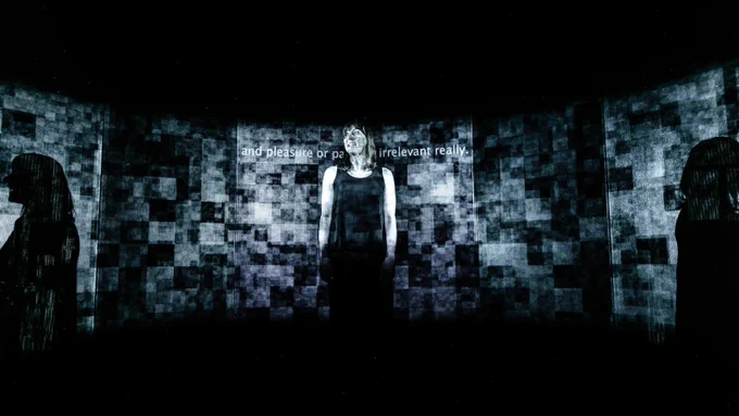 A person stands on a darkened stage in front of a video projection of pixelated squares with integrated captions. Photograph by The Other Richard. 