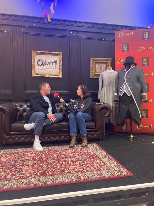 Two people sitting on an old fashioned leather sofa in a set that looks like a Victorian parlour.