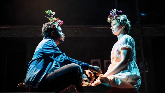 Two people sit opposite one another on a table. Above them is a spotlight shining down. They are both wearing a crown of flowers. Photograph by Alex Brenner. 
