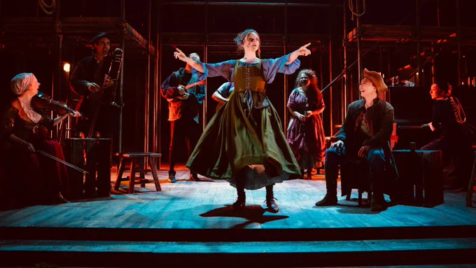 A group of people on stage, playing musical instruments. They stand or sit in a sort of semi-circle around a person in the centre who is pointing out towards the audience. Photograph by Ellie Kurttz. 