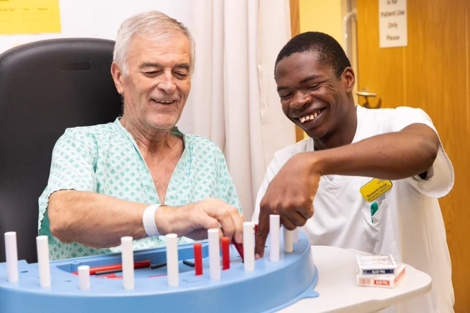 A man in a hospital gown is helped with an activity by a man in a white tunic.