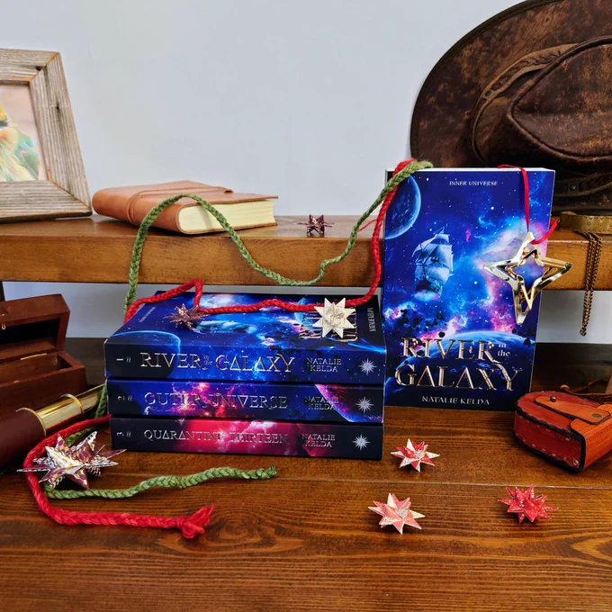 Lifestyle photo of four books on a wooden table with props around them. Three of the books are lying on the side, spine out and are from top to bottom: River in the Galaxy, Outer Universe and Quarantine Thirteen. A copy of River in the Galaxy is also standing upright to the right of the flat laid books. The cover is a blue spacey scene with a wooden ship flying through it and the title in gold across the bottom. The props around and on top of the books lying down are 3d paper stars in reds, white and gold, a green and red braided cord draping around the books and a gold metal star hanging off the book standing up. There is also a leather hat in the back right and a brass spyglass to the left.