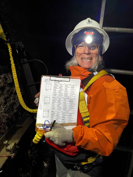 Chela Zabin, wearing a white hard hat, head lamp and an orange jacket, stands at the bottom of a seawall holding a notepad and smiling.
