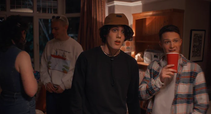 2 people looking forwards in a house, one with a cup in hand the other with a hat on.
