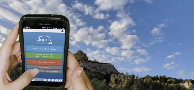 Image of two hands holding up a cell phone with the GLOBE Observer App Clouds Tool on screen. The background contains clouds, trees and mountainous rock.