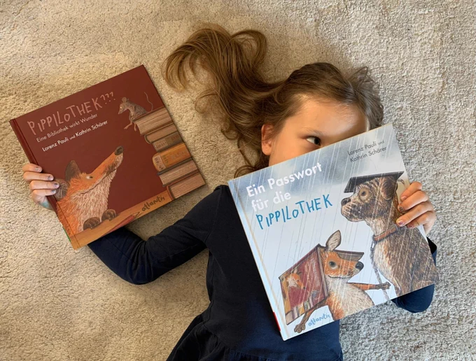 Ein Kind liegt mit dem Rücken auf einem Teppichboden. In der rechten Hand hält es das Kinderbuch "Pippilothek??? Eine Bibliothek wirkt Wunder" vom Körper weg. In der linken Hand hält es den Nachfolgeband "Ein Passwort für die Pippilothek" vor dem Gesicht. (© Foto: Privat)