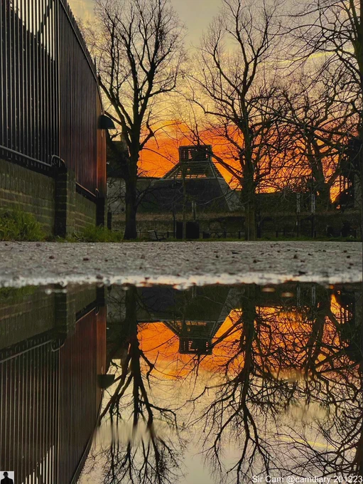 Sunrise in Cambridge, 20 December 2023. Bradley Court building at Darwin College reflected in a puddle with a strong orange sunrise in the background and trees framing the structure.