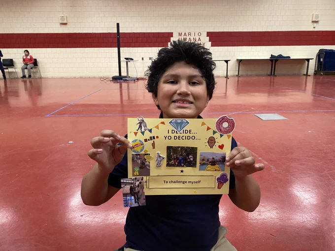 A middle school student sitting on the floor holding up a project he worked on that says “I decide/Yo Decido to challenge myself” with photos pasted on the paper of him kayaking and rock climbing on past Waypoint programs. He has a big smile on his face.