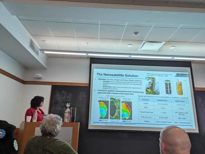 Cadence by a podium giving her thesis defense talk. On the podium she has a model 3U CubeSat (small satellite) next to a loaf of bread to show they are the same size. She is presenting a slide titled “The Nanosatellite Solution”
