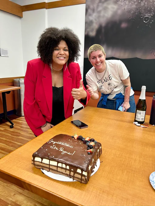 Cadence and one other person smiling posing with a thumbs up behind a chocolate cake that says Congratulations Dr. Payne on it
