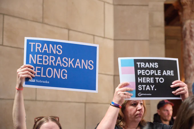 Two people hold ACLU and OutNebraska signs in the Nebraska State Capitol rotunda. The ACLU sign reads "Trans Nebraskans belong" and the OutNebraska sign reads "Trans people are here to stay."