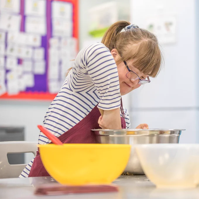 Young girl with Down's Syndrome in a blue and white stripy t-shirt and red apron. She's  wearing glasses with her hair up in a hair tie. She's mixing ingredients in a stainless steel bowl.