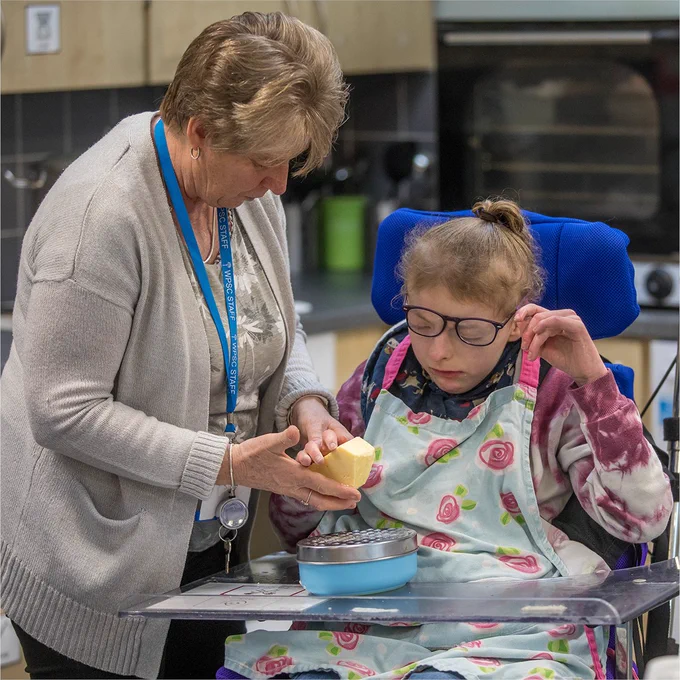 Young girl with Down's Syndrome in a blue and white stripy t-shirt and red apron. She's  wearing glasses with her hair up in a hair tie. She's mixing ingredients in a stainless steel bowl.