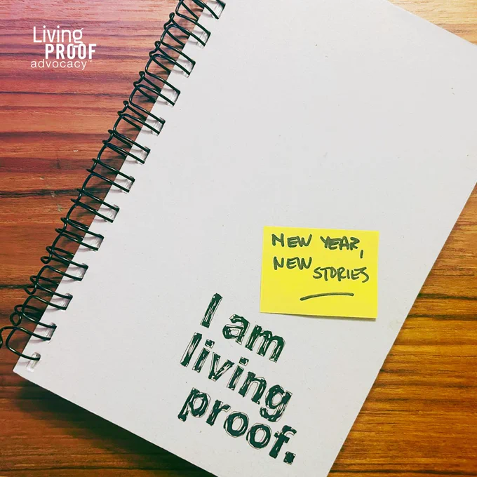 A photo of a spiral-bound notebook with the words I Am Living Proof printed on it. A handwritten sticky note reads "New Year, New Stories."