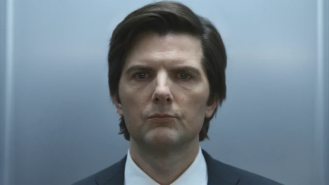 From Severance. Mark Scout (Adam Scott) in a close-up shot, looking straight ahead against a blank white wall. The top of his black suit jacket and white button-down shirt are visible. He wears a miserable expression.
