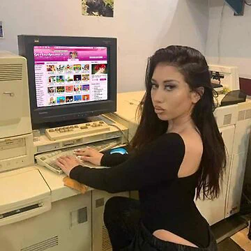 A young femme person looks over their shoulder at the camera as they type on a 1990s desktop