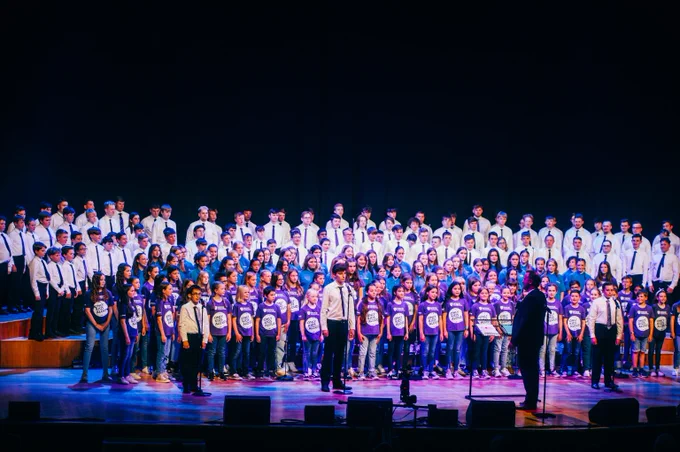 Our Only Boys Aloud, Only Girls Aloud, and Only Kids Aloud choir members perform as a mass choir on the stage at St David's Hall. Three soloists from Only Boys Aloud stand in front of the group.