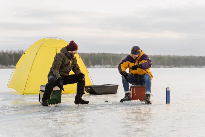 Two men sitting on coolers and ice fishing with a tent behind them. 