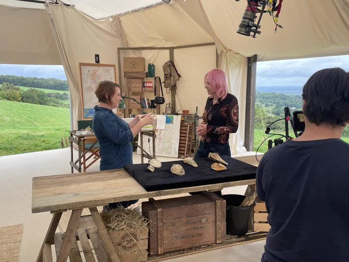Two women talk animatedly behind a table displaying hand axes on a dark cloth. They are standing in a large white canvas tent; the open door flaps reveal a beautiful rolling green landscape. A TV camera operator films their conversation.