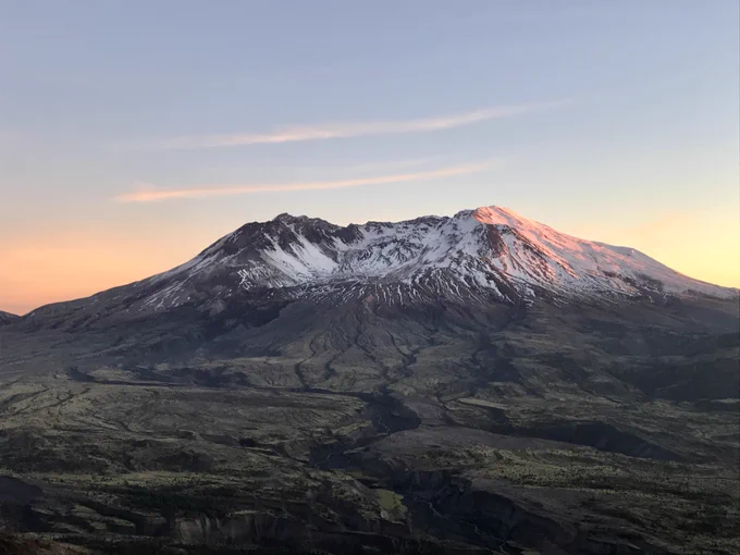 This picture shows a sunset sky and Mount St Helens in the just covered with snow on the top half. The valley below in a green moss color. 