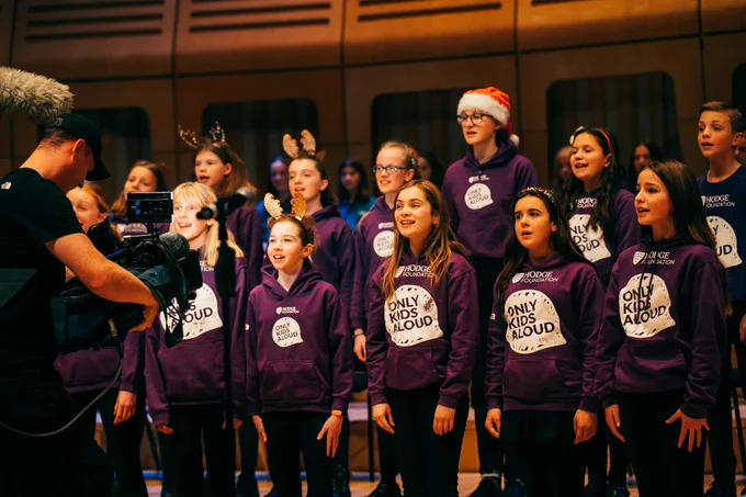Only Kids Aloud members are standing in rows singing. They are wearing purple jumpers and Christmas accessories.
