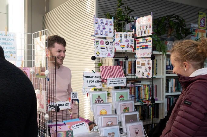 Alex is smiling as someone visits their stall of queer Christmas themed cards.