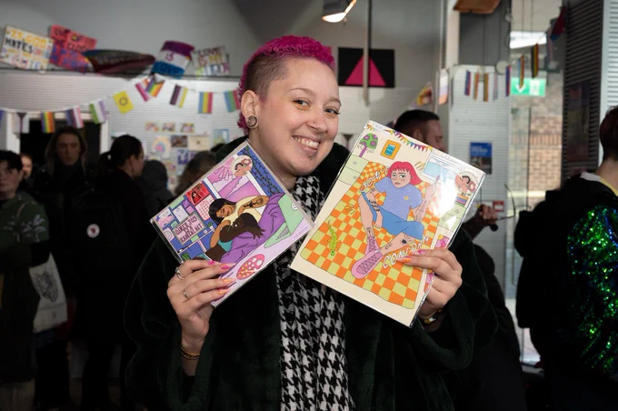 A Winter Market visitor with pink hair is smiling and holding up prints from Boe Studios stall.