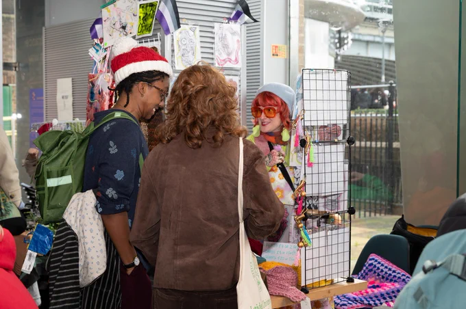 Two visitors to the Winter Market, one of whom is wearing a Christmas hat, are at Sarah-Jane's stall smiling.