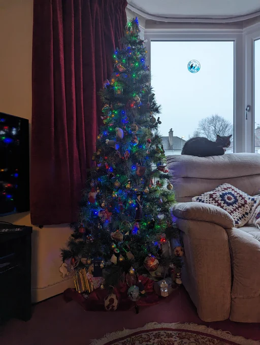 decorated xmas tree with presents under it, next to an armchair with black guard cat perched on the back of it