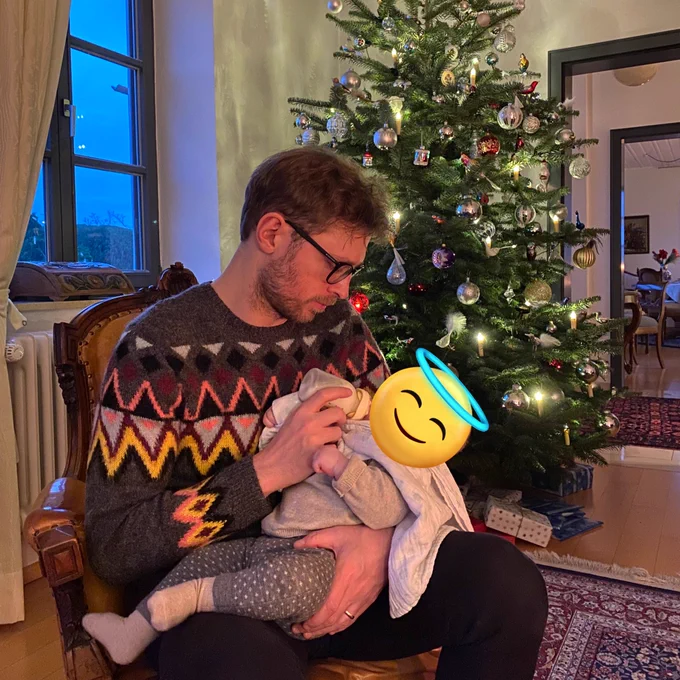 Mann mit schickem Pulli gibt kleinen Engel ein Fläschchen, sitzend vor Weihnachtsbaum.