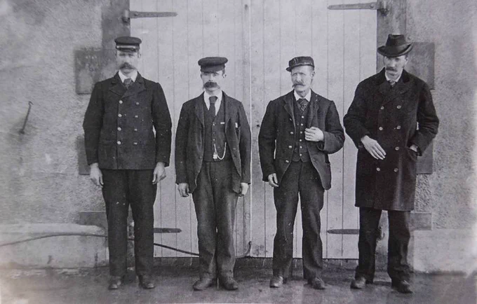 The lightkeepers of Flannan Isles, circa 1900. Four men with moustaches dressed in Victorian uniforms, standing in front of a wooden door. From left to right: James Ducat, Thomas Marshall and Donald McArthur. Superintendent Robert Muirhead, who was on sick leave when his three colleagues disappeared, stands to their right.