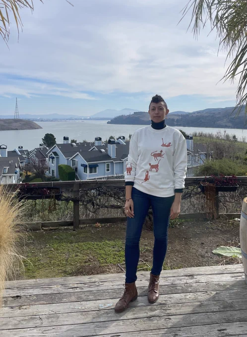 christine facing the camera with a bay in the background. christine is wearing a white crewneck with orange cats on the front and one sleeve.