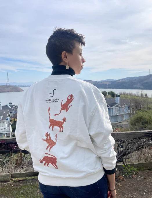 christine with her back facing the camera, wearing a crewneck sweater with four orange cats visible on the back. A logo saying “roots studio” is visible with a subtitle saying “the future is rurban”. A bay is visible beyond christine