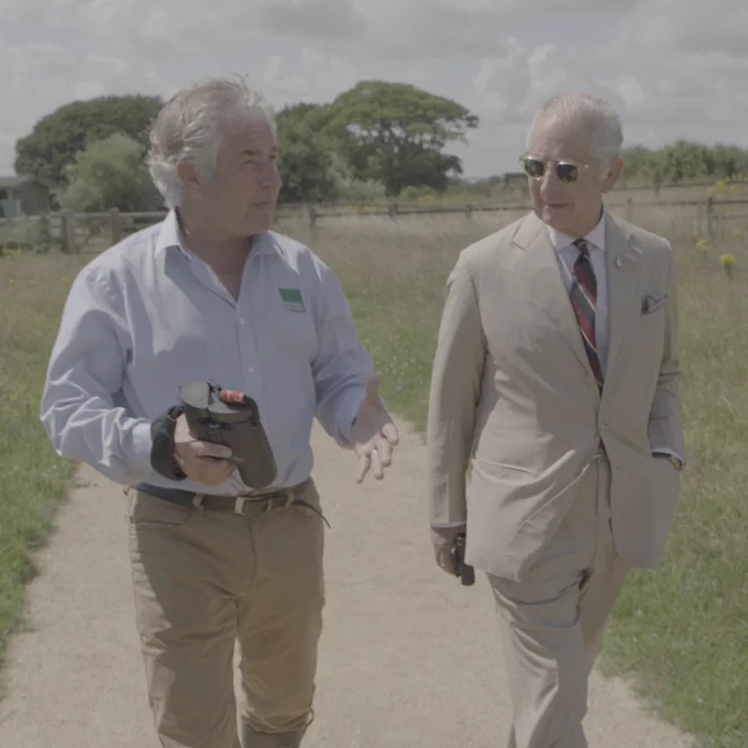 King Charles III and Tony Juniper walk along a path inside the Lincolnshire Coronation Coast National Nature Reserve. It is a summer day and there is long green grass on either side of the path. Tony Juniper is holding binoculars. The King is wearing a light coloured suit and sunglasses.