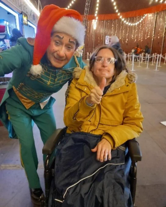Lucy has long brown hair and glasses and wears a mustard yellow jacket. She is a wheelchair user. She has a big thumbs up in the air as there is an elf to the left of her posing for the photo as well. The elf has a green outfit on with a Santa hat. 