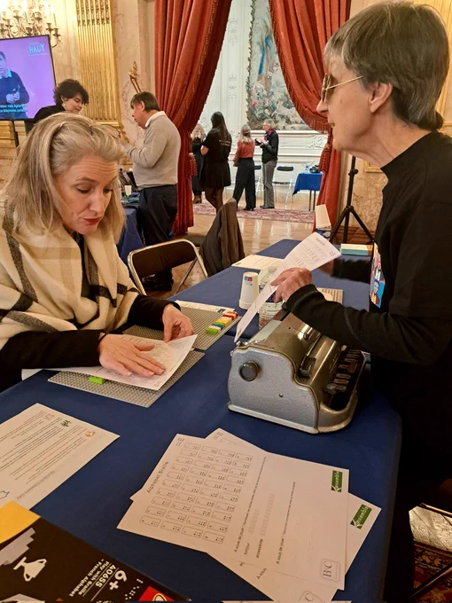 Photo de Servane Hugues à l'assemblée nationale en train de lire une feuille en braille. Elle a les mains posées sur la feuille. Elle est assise à une table bleue.